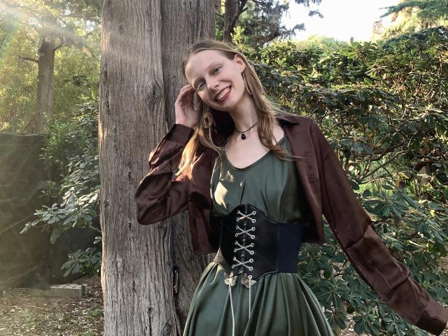 Lallilillo, avec de longs cheveux blonds, une robe verte, un corset noir, une veste marron, souriante, debout près d'un arbre, une main dans les cheveux, ensoleillement, feuilles vertes, un collier noir, mignonne, joli visage..