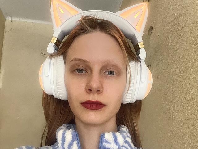 Lallilillo a la peau pâle, du rouge à lèvres, et porte des écouteurs blancs en forme d'oreilles de chat avec des accents orange, ainsi qu'une veste bleue et blanche duveteuse. Elle regarde directement la caméra avec une expression sérieuse..