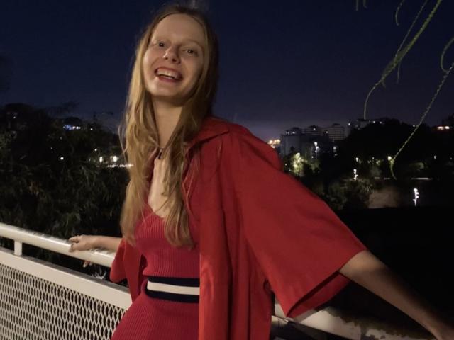 Lallilillo with long blonde hair, red dress, and red graduation cap, smiling at night, leaning on white railing, city lights in background, holding dress with right hand.