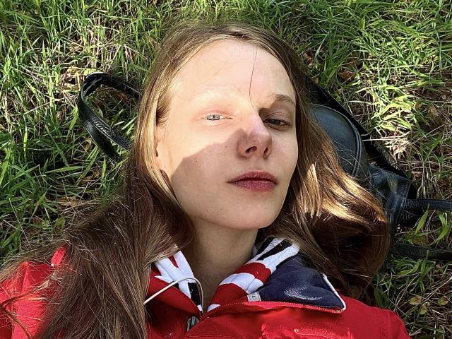 Lallilillo's face is in a photo, lying on grass with sun shadows. She has light brown hair, green eyes, and a red jacket with a Union Jack collar.