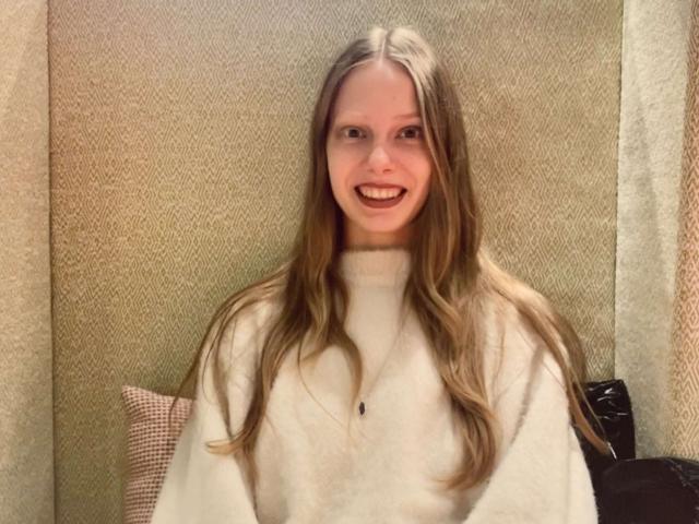 Lallilillo, avec de longs cheveux blonds et lisses, souriante, portant un pull blanc à col roulé, assise contre un mur beige texturé, ayant l'air heureuse et détendue..