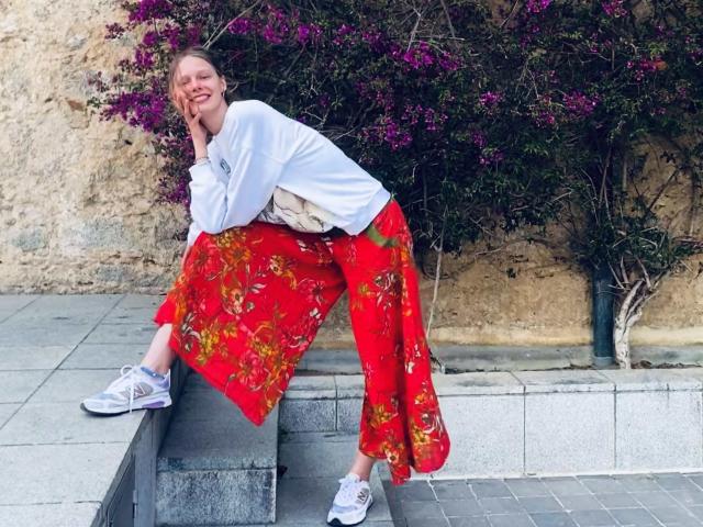 Lallilillo with blonde hair, white long-sleeve shirt, red floral skirt, and white sneakers, sitting on stone ledge, smiling, hand on cheek, purple flowers in background.