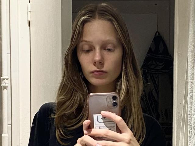 Lallilillo with brown, wavy hair, serious expression, holding a phone, wearing a black shirt, taking a mirror selfie. Her phone has a white sticker on the back.