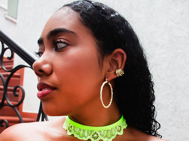 AishaRhodes has dark brown skin, curly black hair, and wears a neon green lace choker with gold hoop earrings and gold ring earrings, looking to the side with red lipstick.