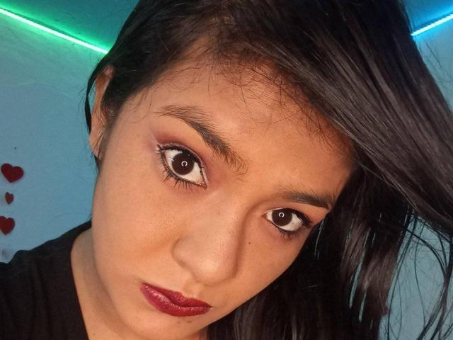 ArianithaXox hat große, braune Augen, dunkel glänzlichen Lippenstift und schwarzes Haar mit einer Seitenteilung. Sie blickt ernst drein, ihr Gesicht ist leicht geneigt..