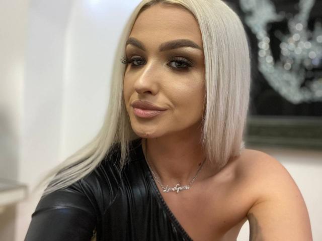 KylieJonnes, con lunghi capelli biondi, sopracciglia scure e una maglietta nera con una spalla scoperta, mostra un sorriso appena accennato con una piccola goccia di liquido sul mento..