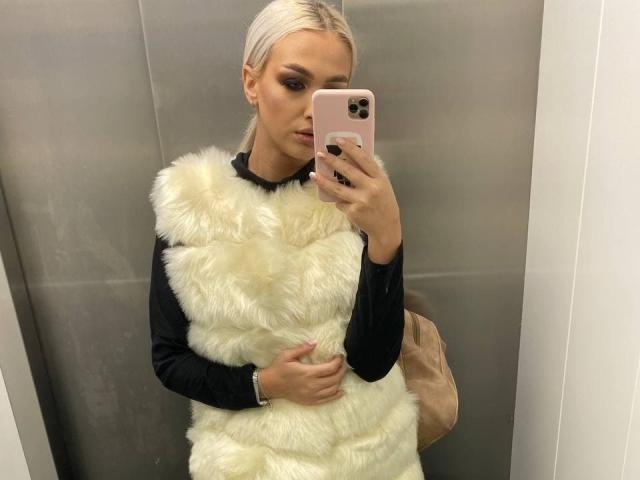 KylieJonnes, con i capelli biondo platino, indossa un gilet bianco soffice sopra una maglia nera a maniche lunghe, e sta scattando un selfie allo specchio in un ascensore, tenendo in mano un iPhone rosa..