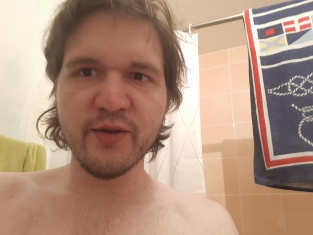 NectarineFad is a shirtless, slightly chubby guy with brown, wavy hair, a beard, and a light skin tone, standing in a bathroom with a towel hanging.