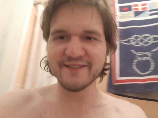 Shirtless NectarineFad with wavy brown hair, beard, and slight smile, showing a hint of chest hair, in a blurry selfie.
