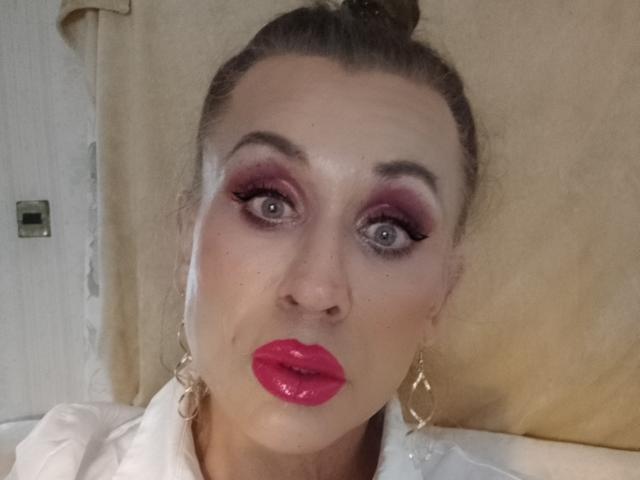 LilianaShe has pink lipstick, red eyeshadow, and gold hoop earrings. She's looking surprised with wide blue eyes and a raised brow. Her brown hair is in a bun.
