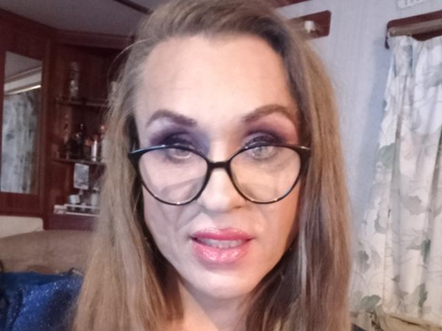 LilianaShe has glasses, light brown hair, purple eyeshadow, pink lipstick, and a slight smile, showing her pouty ass-kissing lips. Her tits are slightly visible under her blue shirt.