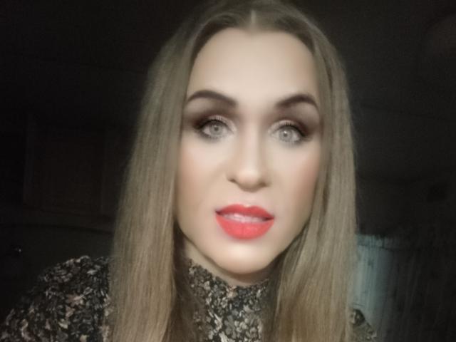 LilianaShe has long blonde hair, light makeup, red lipstick, and a subtle smile. She's wearing a dark, textured top. Her eyes are light blue, and she's looking at the camera.