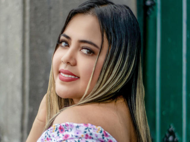 GraceJansen with blonde ombre hair, smiling with pink lips, brown eyes, and wearing a floral off-shoulder top, looking over her shoulder. Her tits are partially visible.