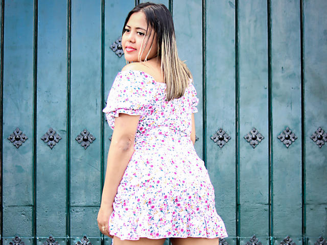 GraceJansen has medium brown skin, black and blonde ombre hair, and wears a floral off-shoulder dress. She's looking over her shoulder with a slight smile.