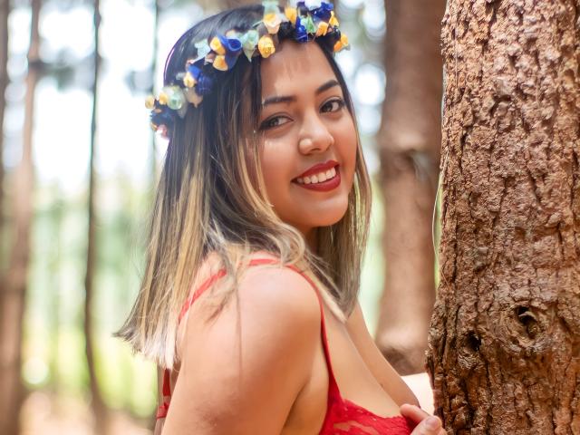 GraceJansen with blonde ombre hair, red lace bra, smiling, wearing a blue-yellow flower crown, standing against a tree, showing her tits, in a forest.