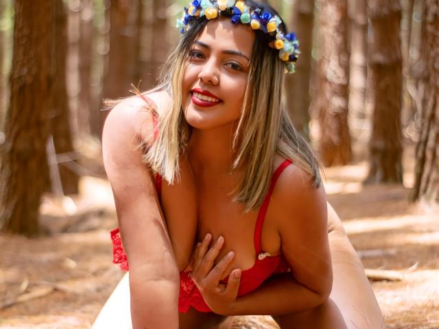 GraceJansen with blonde hair, red dress, and flower crown, leaning forward, hand on her tits, smiling, forest background, sunlit, red lipstick, sexy, cute, attractive.