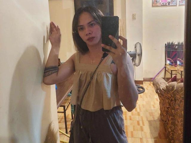 SuckableMistress - uma mulher atraente com cabelo preto na altura dos ombros, vestindo uma blusa bege e calças cinzas escuras, segurando um telefone, em pé contra uma parede, mostrando uma tatuagem no braço direito..