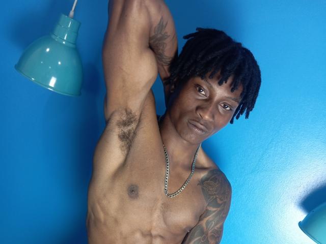 Tachala, a muscular black man with dreadlocks, raises his arm to show hairy armpits. He's shirtless, wearing a chain, with tattoos on his right arm. Blue wall.