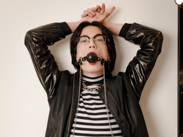 MilarHole with arms raised, black leather jacket, striped shirt, ball gag in mouth, chain around neck, glasses, black hair, white wall, standing, looking up, slightly parted lips.