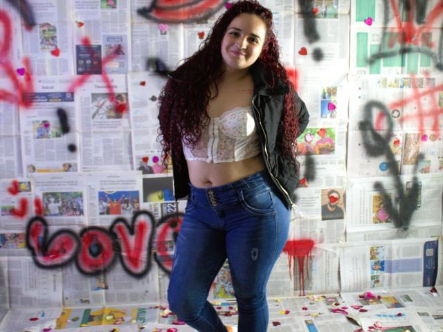 LunaF with curly red hair, smiling, wearing a floral lace bra, black jacket, and blue jeans, standing in a graffiti-covered room with red and black spray paint.