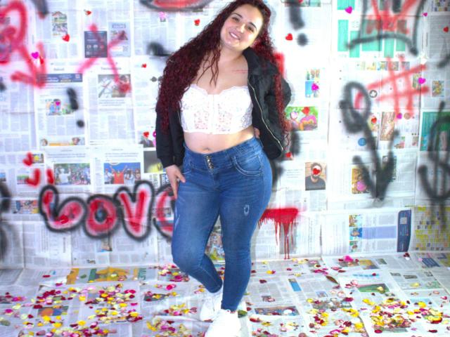 LunaF with curly red hair, white lace bra, black jacket, blue jeans, white sneakers, standing, smiling, hands in pockets, colorful graffiti, newspaper wall, confetti on floor.