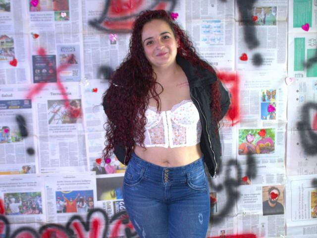LunaF with curly red hair, white floral bra, black jacket, and blue jeans, standing with hands behind back, smiling, wearing a small tattoo on her right chest.