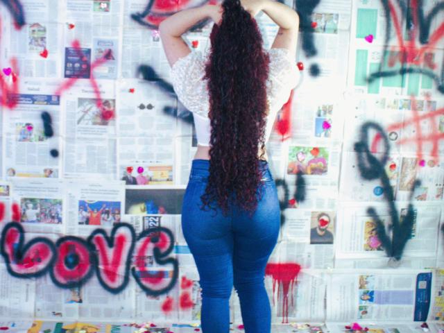 LunaF with long curly red hair, wearing a white sparkly top and tight blue jeans, standing with hands behind head, facing graffiti-covered wall. Boobs not visible, ass prominent.