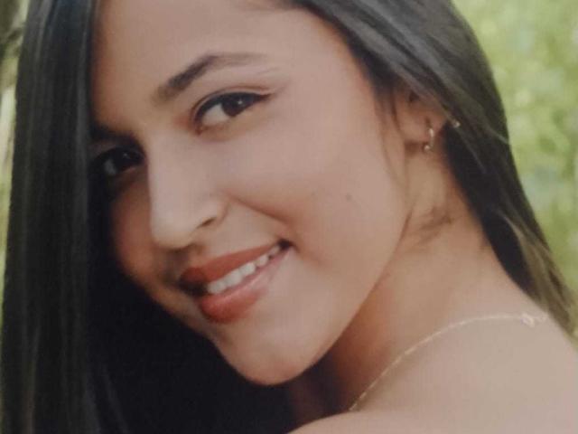 HikariChloe has smooth brown skin, long black hair, and is smiling with red lips showing. She has small tits and is wearing a gold necklace with a tiny cross.
