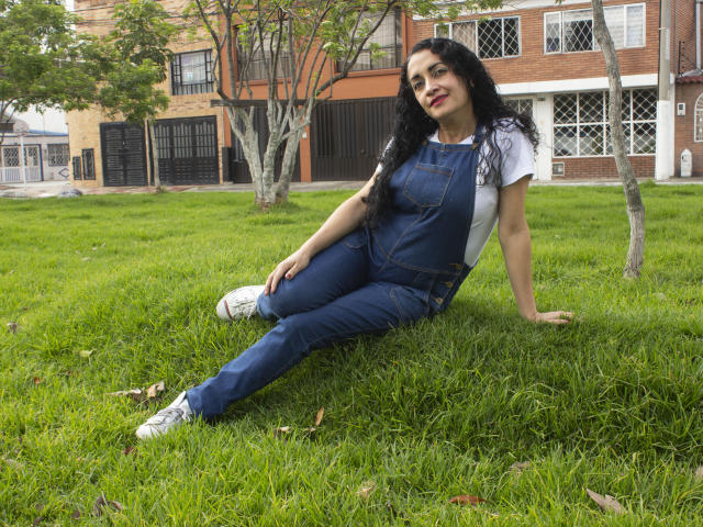 TitaNaughty met lang zwart haar, gekleed in een denim overall, een wit T-shirt en witte sneakers, zittend op groen gras, leunend achterover met een glimlach, waarbij haar achterwerk zichtbaar is..