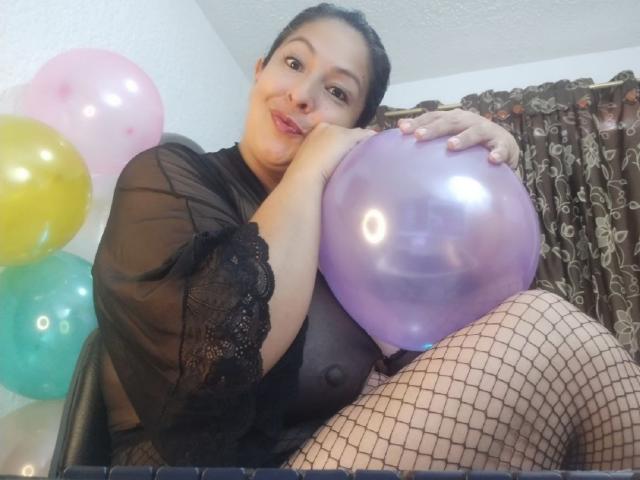 CatalinaJH with dark hair, black lace top, fishnet stockings, holding purple balloon, sitting, smiling, showing bare tits, colorful balloons in background.