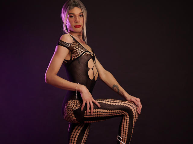 ElyassDick, com cabelo loiro liso, vestindo um macacão preto de rede de peixe com recortes, expondo seios e vulva, tatuagem no braço direito, joelho levantado, iluminação roxa e preta..