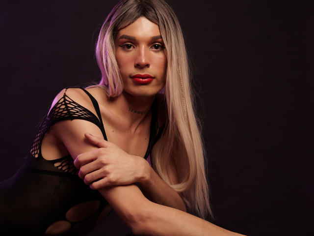 ElyassDick, con lunghi capelli biondi, indossa una maglia nera a rete, ha il rossetto rosso ed è piegato in avanti con le braccia incrociate, mostrando piccoli seni e una figura snella e sexy..