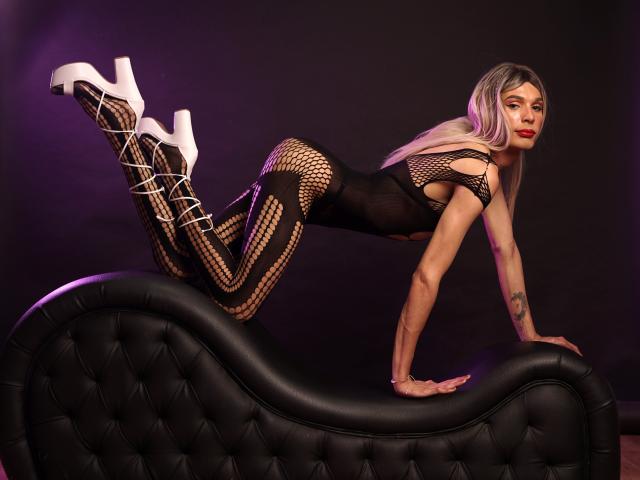 ElyassDick con un body de malla negra, tacones blancos de plataforma, sentado en un sofá negro con capitoné, con las rodillas levantadas y las manos abajo, mirando a la cámara, mostrando el trasero, con un tatuaje en el antebrazo derecho, y cabello rubio largo..
