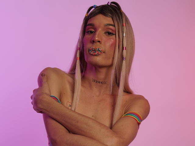 ElyassDick com maria-chiquinhas loiras, contas coloridas no cabelo, rosquinha colorida na boca, braços cruzados, sem camisa, braceletes coloridos, tatuagem no pescoço, iluminação rosa-roxo..