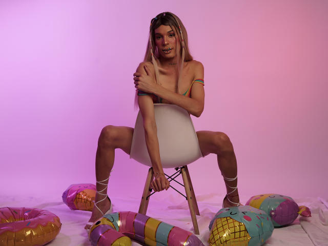 ElyassDick, sem camisa, sentado em uma cadeira branca com as pernas abertas, cobrindo os pequenos seios com o braço, usando uma braçadeira colorida, sapatos brancos com cadarço, cercado por donuts infláveis coloridos, iluminação rosa-roxo..