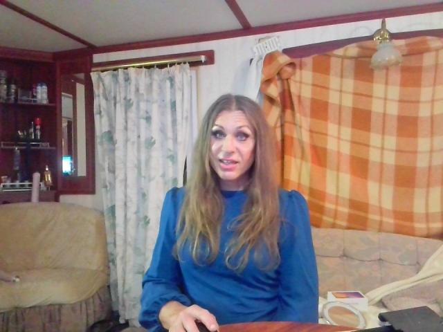 LilianaShe has long brown hair, a blue long-sleeve shirt, and a slightly open mouth with a hint of a smile, sitting in a cozy room with curtains.