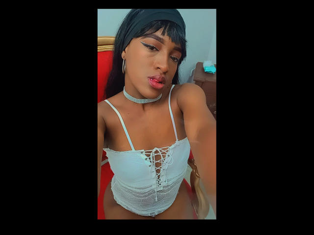 LilaJamez with dark skin, black headband, white lace-up top, choker, pink lips, and hoop earrings, showing off her small tits and sexy look in a selfie.