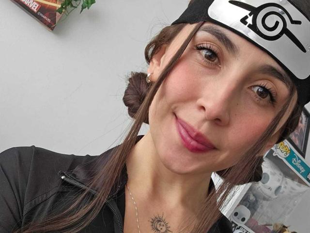 Rossy Nolen tiene una cara linda, ojos marrones, lápiz labial rosa y un pequeño tatuaje de un sol con rayos en su pecho. Lleva una diadema negra y una chaqueta negra..