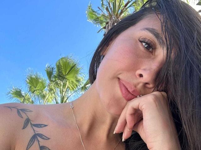 RossyNolen has a tattoo on her shoulder, brown hair, and is looking at the camera with a slight smile. She's wearing a necklace, and her ass isn't visible.