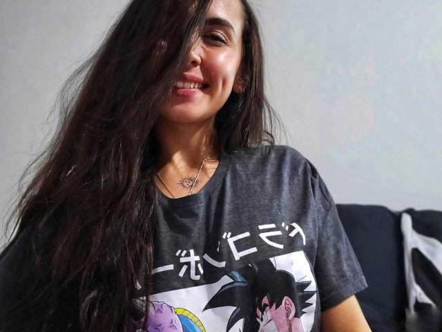 RossyNolen with long, dark, messy hair, smiling, wearing a grey Dragon Ball Z t-shirt, showing teeth, and a small necklace, standing in front of a dark couch.
