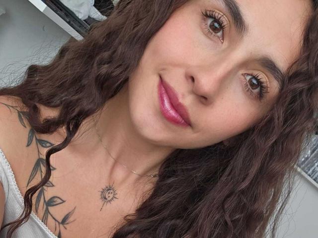 Rossy Nolen, con cabello castaño rizado, lápiz labial rosa y un tatuaje de sol en el pecho, vestida con una blusa blanca con un hombro descubierto, sonriendo suavemente a la cámara..