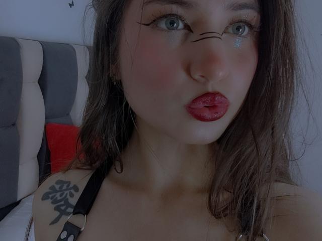 SopphiesWett has a tattoo on her shoulder, pouty red lips, and glittery blue eyes with black eyeliner, wearing a strap, in a dimly lit bedroom.