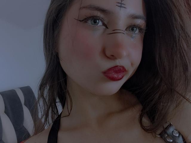 SopphiesWett with dark brown hair, blue eyes, black eyeliner, red lipstick, and a small tattoo above her right eyebrow. She has a cute, pouty expression. Boobs visible in a black top.