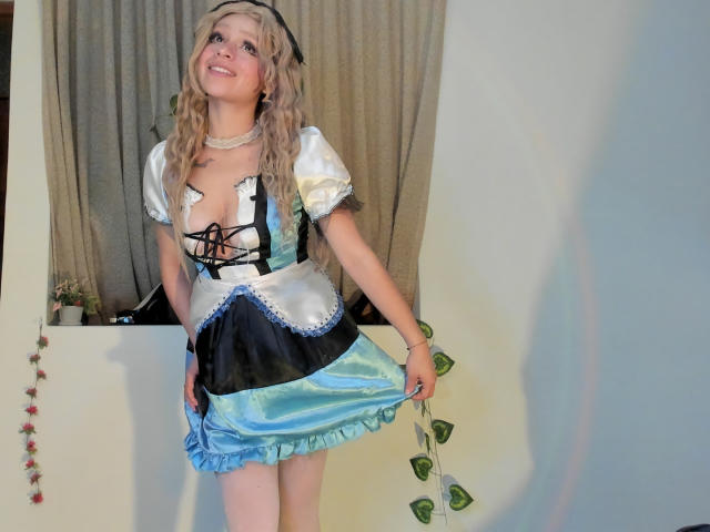 Blonde Hesttia in a French maid outfit, lifting her blue and black skirt, showing her tits, smiling with curly hair, wearing a black headband and necklace, in a room.