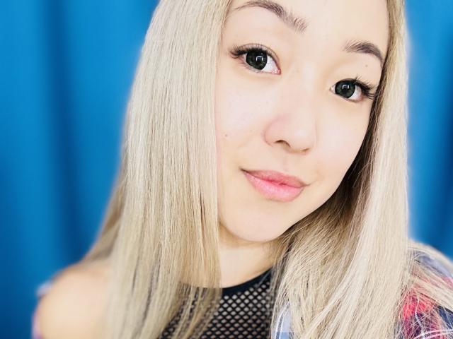 RenyLime has blonde hair, green eyes, and pink lips. She's wearing a black mesh top, looking directly at the camera with a slight smile. Her ass is not visible.