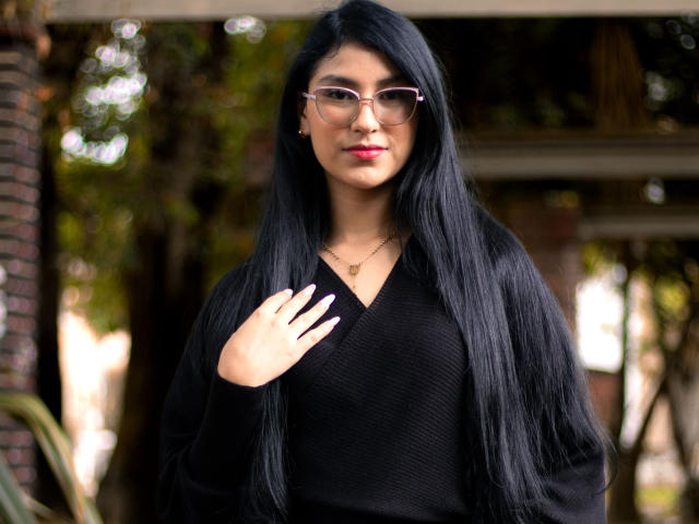 Ema Henderson mit langen, schwarzen Haaren, rosa Brille, rotem Lippenstift, einem schwarzen Pullover mit V-Ausschnitt und einer goldenen Halskette, die ihren Pullover mit einer Hand hält und draußen steht..