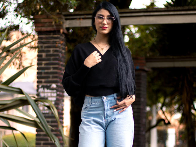 Ema Henderson mit langen, schwarzen Haaren, Brille, schwarzem Pullover und hellblauen Jeans, steht draußen, eine Hand auf dem Pullover, die andere auf der Hüfte, blickt in die Kamera und präsentiert ihr Hinterteil..