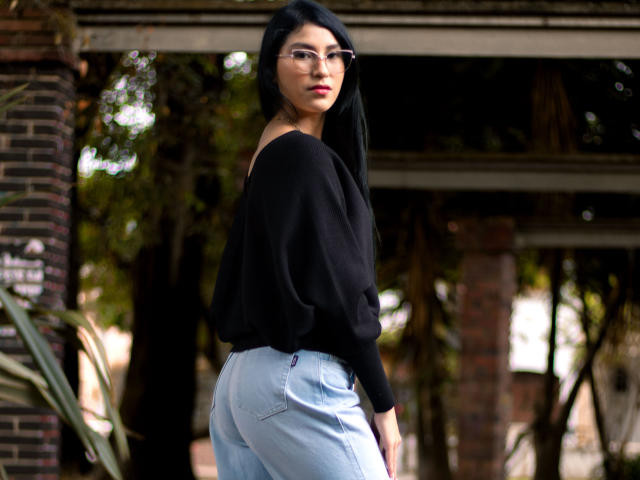 Ema Henderson, com cabelos longos e pretos, uma blusa preta com um ombro de fora e jeans azul claro, mostrando suas nádegas arredondadas, está em pé do lado de fora, olhando para trás com uma expressão séria..