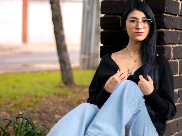 Ema Henderson mit langem, schwarzem Haar, Brille und pinkem Lippenstift, sitzt an einer Ziegelwand, trägt ein schwarzes Hemd und hellblaue Jeans und hält ihr Hemd..