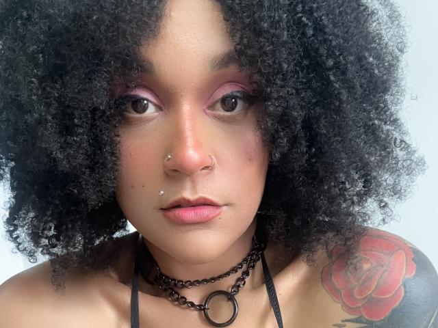AmyStone69 with curly black hair, nose piercings, pink eyeshadow, black chain necklace, and a red rose tattoo on her right shoulder, staring directly at the camera.